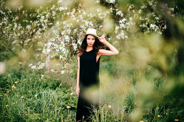 Young adorable innocent brunette girl in black dress in spring blooming garden posing for camera. female portrait outdoor in park. Blossom flowers on trees. Young unusual person enjoying nature