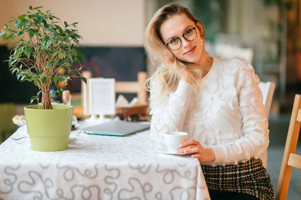 Young beautiful blonde lady in white sweater and glasses sitting in caffe and posing for camera with positive face expression.