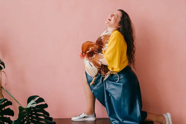 Young beautiful long haired excited girl hugs chicken and laughing on pink background. Lifestyle portrait of eccentric woman with funny expressive emotional face holding rooster in her hands