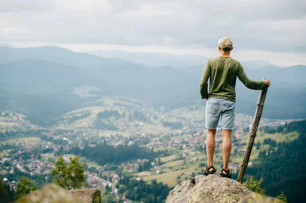 Lifestyle summer portrait from behind of successful man with wooden stick standing on top of mountaing with beautiful landscape in front. Male traveler enjoying nature view from highest peak at hill