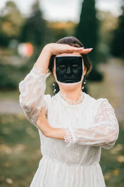 夏の公園で彼女の手でジェスチャーを示す創造的な顔の芸術 黒の正方形 と結婚式のドレスの奇妙な女性 — ストック写真