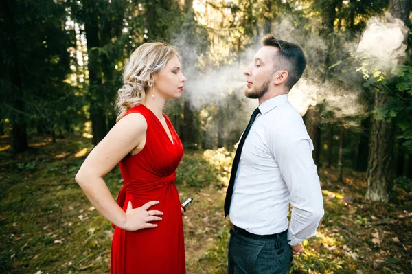 Odd excited happy loving couple standing outdoor and blowing thick clouds of vape smoke to each others face at nature. Elegant pair of lovers strange relationship. Addiction from delight of smoking.