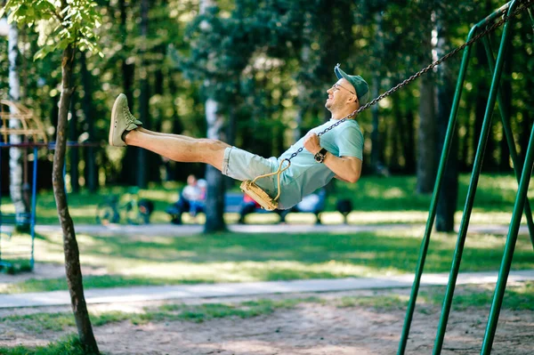 夏に子供の遊び場の都市公園でスイングに乗ってメガネで大人のスタイリッシュな男 幸せな男は子供の頃を覚えている 上下の動きを楽しむ魅力の男性の人 奇妙な男の子の屋外 — ストック写真