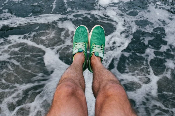 Tanned hairy strong male legs in green summer lightweight sneakers. Unrecognizable man powerful legs with muscles on abstract waterfall in motion background. Masculine relax and meditation concept.