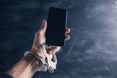 Elleri kelepçeli, gri arka planda bir telefon tutuyor.