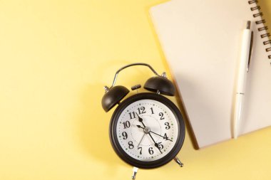 Alarm clock, notebook and pen on yellow background with copy space