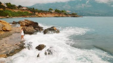 Beyaz elbiseli kadın, Yunanistan 'ın Kefalonia kentinde deniz dalgaları düşerken kayalık sahilde duruyor.
