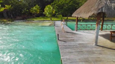 Meksika 'da tropik bir barakanın altında, turkuaz su kenarında tahta iskelede yürüyen bir kadın..