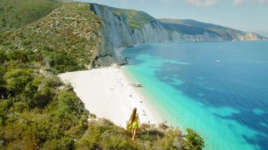 Uçurumdaki kadın, tenha beyaz plajları ve İyon Denizi 'nin turkuaz suyunu zevkli teknelerle ve güneşlenen insanlarla gözden kaçırıyor..