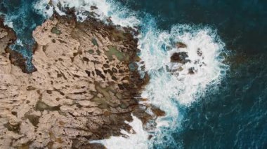 Dalgaların gelgit havuzları ve katmanlı kıyı kayaları üzerinde çarpışını gösteren hava manzarası.