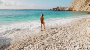 Yunanistan 'da Kefalonya' da turkuaz deniz kenarında çakıl taşı plajında çıplak ayakla yürüyen kadın.