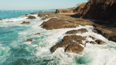 Kayalık sahilde yürüyen kişi açık mavi gökyüzü altında okyanus dalgalarının düşüşünü izliyor..