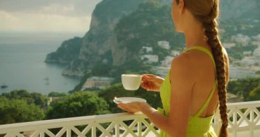 Yaz seyahati sırasında Capri kayalıklarına ve denize bakan bir balkonda sabah kahvesinin keyfini çıkaran bir kadın..