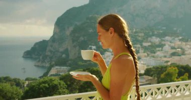 Bir kadın, Capri İtalya 'nın sakin sahil şeridine bakan bir balkonda sabah kahvesinin tadını çıkarıyor..