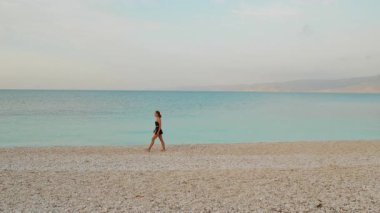 Sakin turkuaz İyon Denizi 'nin yanında, Kefalonia çakıl taşı plajı boyunca yürüyen bir kadın..