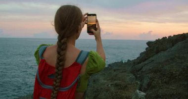 Solo gezgin kayalık sahilde gün batımını akıllı telefonuyla kaydediyor..