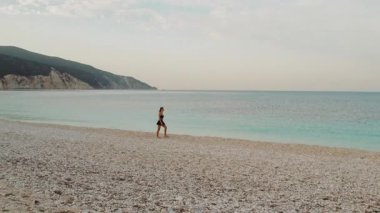 Uzak dağları ve pastel gökyüzü ile sessiz Kefalonia sahillerinde gezinen bir kadın..