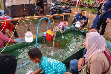 Yogyakarta- 4 Ekim 2025: Çocuklar gece pazarında eğlenceli bir balık tutma yarışmasına katılıyor. Çocuklar basit oltaları kullanarak renkli bir su havuzundan küçük oyuncak balıklar yakalamaya çalışıyorlar.