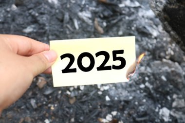 Üzerinde 2025 yazılı, yarım yamalak bir banknot tutuyor. Elveda 2025 kavramı ve yeni yıl kavramı 2026. 2025 kağıt yanıyor, 2025 'e elveda diyor ve 2026' nın vaadini memnuniyetle karşılıyor.