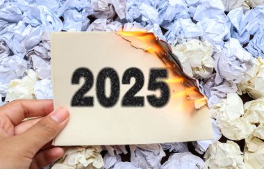 Üzerinde 2025 sayısının yazılı olduğu, buruşuk beyaz kağıtlarla çevrili, kömür dokusu efektiyle yazılmış yarısı yanmış bir kağıt parçasını tutan bir el. Elveda 2025 kavramı ve yeni yıl kavramı 2026. 2025 'in sonu
