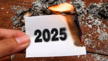 Üzerinde 2025 sayısının yazılı olduğu, buruşuk beyaz kağıtlarla çevrili, kömür dokusu efektiyle yazılmış yarısı yanmış bir kağıt parçasını tutan bir el. Elveda 2025 kavramı ve yeni yıl kavramı 2026. 2025 'in sonu
