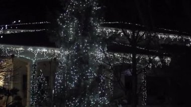 Açık hava Noel ağacı, parlak beyaz LED dizgi ışıkları ve modern ev cephesinde şenlikli sarkık çelenklerle parıldayan tatil süslemeleri, rahat kış atmosferi yaratıyor.