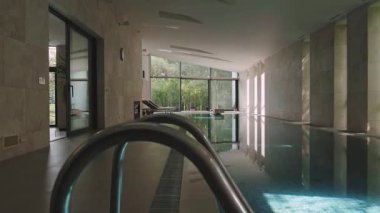 Lüks Kaplıcada Büyük Kapalı Yüzme Havuzu Doğal Taş Duvarları, Panoramik Pencereler, Yumuşak Gün Işığı ve Yeşil Bahçe Görünümü, İnsansız Huzur Sağlığı Videosu