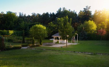 Yeşil ağaçlar, çimenler ve ormanlarla çevrili Beyaz Ahşap Çardaklı Huzurlu Bahçe Manzarası Altın Saat Yaz Akşamı