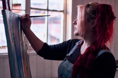 Güzel, kızıl saçlı, boya fırçasıyla poz veren ve stüdyosunda doğal ışık altında resim yapan, çok neşeli, kot pantolon giyen bir Latin. Bağımsız kadın, güzellik ve tavır.