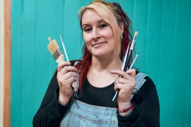 Güzel, neşeli, kızıl, Latin, yetişkin bir kadının portresi. Davranışlarıyla poz veriyor ve kot pantolon giyiyor. Ona stüdyosunda birkaç boya fırçası eşlik ediyor.                               