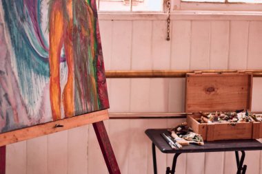 Bir atölyede, doğal ışığı olan bir pencerenin yanında, üzerinde birkaç renkli boya ve fırça bulunan bir kutunun yanında bir resim görüntüsü