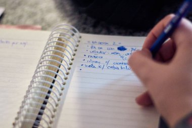 Genç bir kadının elleri, not defterine yazıp not alıyor, detayları, kelimeleri, altçizgileri, renkleri ve kaleminin hareketlerini vurguluyor. Kapalı.