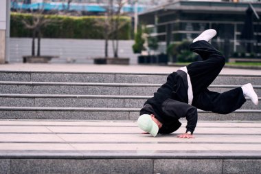 Genç bir adam enerjik bir şekilde break dans yapıyor ve bazı ofis binalarının önünde amuda kalkıyor, rahat kıyafetler giyiyor, bir sürü tavırları, hareketleri ve iyi enerjisi var. Sokak dansı, serbest stil.