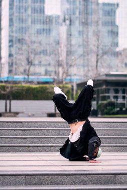 Genç bir adam enerjik bir şekilde break dans yapıyor ve bazı ofis binalarının önünde amuda kalkıyor, rahat kıyafetler giyiyor, bir sürü tavırları, hareketleri ve iyi enerjisi var. Sokak dansı, serbest stil.