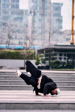 Genç bir adam enerjik bir şekilde break dans yapıyor ve bazı ofis binalarının önünde amuda kalkıyor, rahat kıyafetler giyiyor, bir sürü tavırları, hareketleri ve iyi enerjisi var. Sokak dansı, serbest stil.