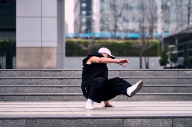 Genç bir adam enerjik bir şekilde break dans yapıyor ve bazı ofis binalarının önünde amuda kalkıyor, rahat kıyafetler giyiyor, bir sürü tavırları, hareketleri ve iyi enerjisi var. Sokak dansı, serbest stil.