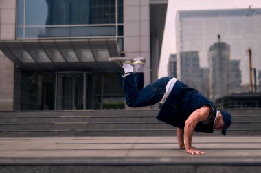 Genç, zinde bir yetişkin coşkuyla break dans yapıyor, kollarını açıyor, şınav çekiyor ve ritme ayak uyduruyor. Kot pantolon, şehir kıyafeti ve kep giymişti. Tavır, hareket ve enerji dolu. Sokak dansı, serbest stil, modern dans..