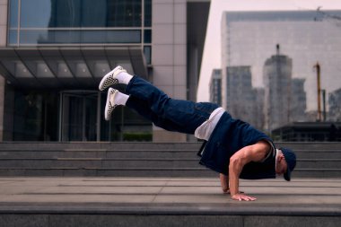 Genç, zinde bir yetişkin coşkuyla break dans yapıyor, kollarını açıyor, şınav çekiyor ve ritme ayak uyduruyor. Kot pantolon, şehir kıyafeti ve kep giymişti. Tavır, hareket ve enerji dolu. Sokak dansı, serbest stil, modern dans..