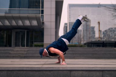 Genç, zinde bir yetişkin coşkuyla break dans yapıyor, kollarını açıyor, şınav çekiyor ve ritme ayak uyduruyor. Kot pantolon, şehir kıyafeti ve kep giymişti. Tavır, hareket ve enerji dolu. Sokak dansı, serbest stil, modern dans..