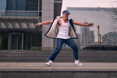 Genç, zinde bir yetişkin coşkuyla break dans yapıyor, kollarını açıyor, şınav çekiyor ve ritme ayak uyduruyor. Kot pantolon, şehir kıyafeti ve kep giymişti. Tavır, hareket ve enerji dolu. Sokak dansı, serbest stil, modern dans..