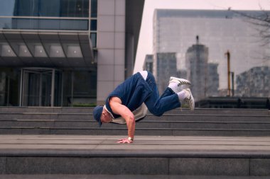 Genç, zinde bir yetişkin coşkuyla break dans yapıyor, kollarını açıyor, şınav çekiyor ve ritme ayak uyduruyor. Kot pantolon, şehir kıyafeti ve kep giymişti. Tavır, hareket ve enerji dolu. Sokak dansı, serbest stil, modern dans..