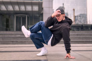 Genç ve zinde bir yetişkin kameraya poz vererek break dans yapıyor. Şehir kıyafeti ve kep giymişti. Bir sürü tavır, hareket ve iyi enerji. Sokak dansı, serbest stil, modern dans..