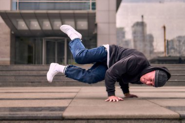 Genç, zinde bir yetişkin coşkuyla break dans yapıyor, amuda kalkıyor ve kendini kaldırıyor. Kot pantolon, şehir kıyafeti ve kep giymişti. Bir sürü tavır, hareket ve iyi enerji. Sokak dansı, serbest stil, modern dans..