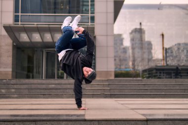 Genç, zinde bir yetişkin coşkuyla break dans yapıyor, amuda kalkıyor ve kendini kaldırıyor. Kot pantolon, şehir kıyafeti ve kep giymişti. Bir sürü tavır, hareket ve iyi enerji. Sokak dansı, serbest stil, modern dans..