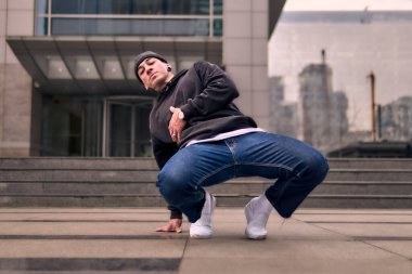 Genç ve zinde bir yetişkin kameraya poz vererek break dans yapıyor. Şehir kıyafeti ve kep giymişti. Bir sürü tavır, hareket ve iyi enerji. Sokak dansı, serbest stil, modern dans..