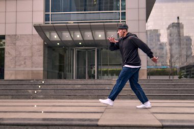 Formda bir genç adam coşkuyla break dans yapar, sert adam pozları verir, şınav çeker ve ritme ayak uydurur. Kot pantolon, sokak kıyafeti ve şapka giyiyor. Tavır, hareket ve enerji dolu. Sokak dansı, serbest stil ve modern dans..