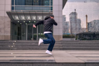 Formda bir genç adam coşkuyla break dans yapar, sert adam pozları verir, şınav çeker ve ritme ayak uydurur. Kot pantolon, sokak kıyafeti ve şapka giyiyor. Tavır, hareket ve enerji dolu. Sokak dansı, serbest stil ve modern dans..