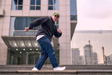 Formda bir genç adam coşkuyla break dans yapar, sert adam pozları verir, şınav çeker ve ritme ayak uydurur. Kot pantolon, sokak kıyafeti ve şapka giyiyor. Tavır, hareket ve enerji dolu. Sokak dansı, serbest stil ve modern dans..