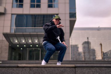 Genç ve zinde bir yetişkin kameraya poz vererek break dans yapıyor. Şehir kıyafeti ve kep giymişti. Bir sürü tavır, hareket ve iyi enerji. Sokak dansı, serbest stil, modern dans..