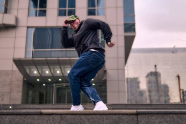 Formda bir genç adam coşkuyla break dans yapar, sert adam pozları verir, şınav çeker ve ritme ayak uydurur. Kot pantolon, sokak kıyafeti ve şapka giyiyor. Tavır, hareket ve enerji dolu. Sokak dansı, serbest stil ve modern dans..